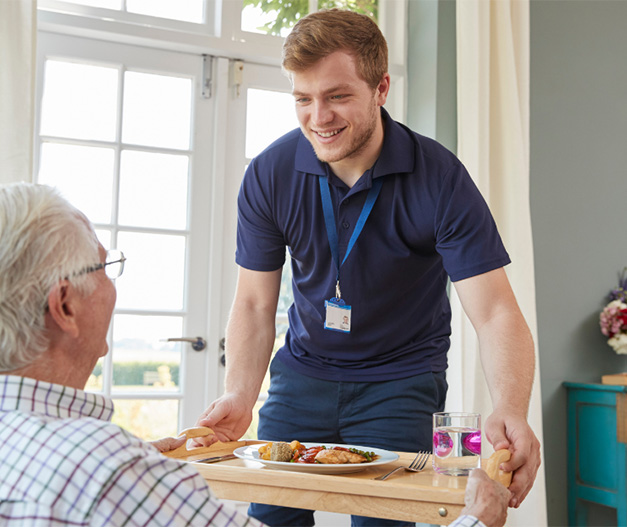 A young male care assistant bringing an elderly gentleman his dinner