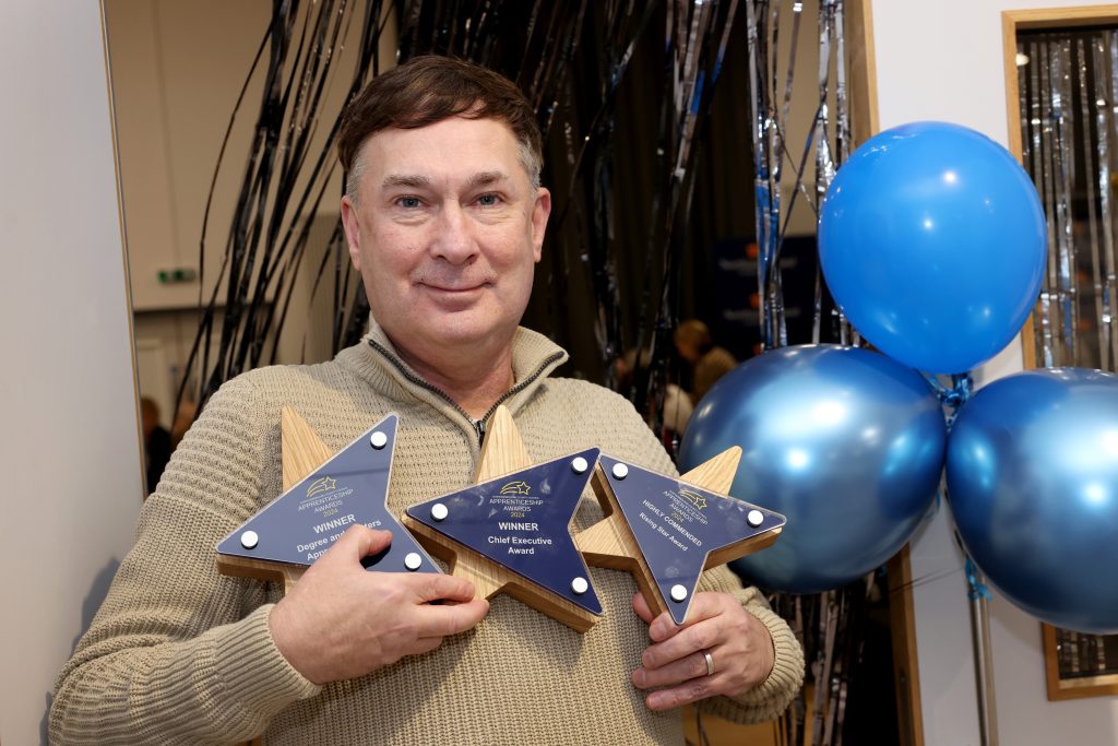 Man smiling at the camera while holding three award trophies. Balloons and streamers are in the background.